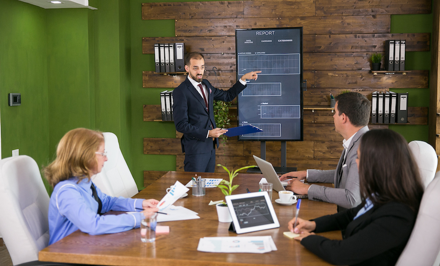 Businessman in suit pointing at charts on tv screen in a meeting with his colleagues. Leadership meeting in conference room.